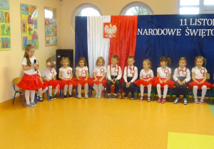 Dziewczynka trzyma w ręku mikrofon, recytuje wiersz. Grupa dzieci po prawej stronie siedzi na krzesełkach. W tle tablice udekorowane granatowym materiałem z godłem Polski.